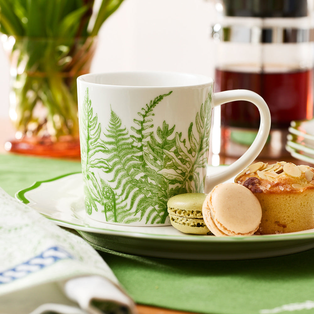 A Caskata Spring Verde Mug, decorated with green fern designs, rests on a plate alongside two macarons and a pastry, while a French press and flowers add brightness to the scene.