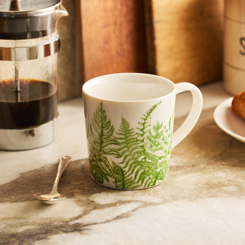 The Caskata Spring Verde Mug, featuring green fern patterns, rests on a countertop beside a silver spoon, French press, and a plate holding a croissant.
