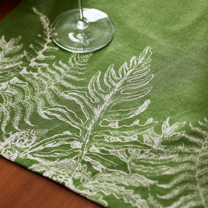 A clear wine glass sits on Caskata’s Spring Verde Table Runner, its green fabric adorned with white fern patterns for a charming cottage-core look.