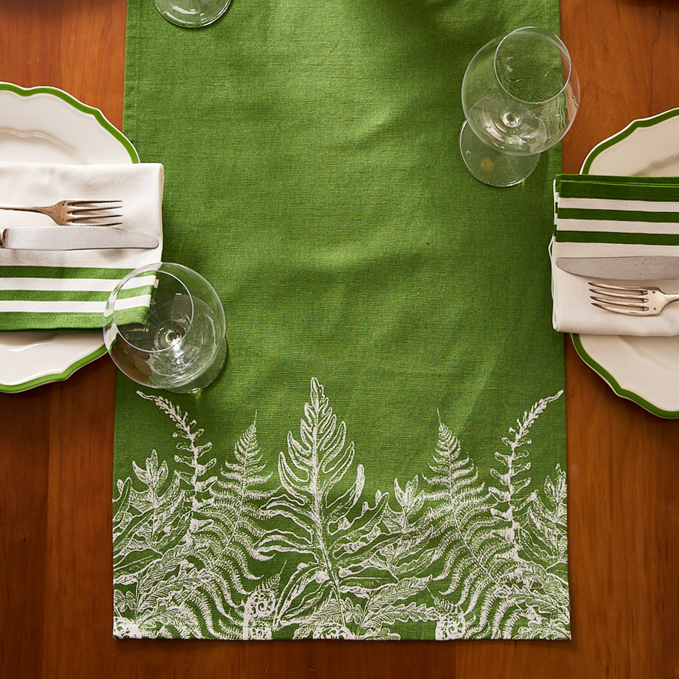 Two place settings with green and white striped napkins on white plates, arranged around a wooden table featuring the Caskata Spring Verde Table Runner, and empty wine glasses—perfect for a cozy cottage-core style.