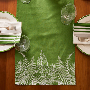 Two place settings with green and white striped napkins on white plates, arranged around a wooden table featuring the Caskata Spring Verde Table Runner, and empty wine glasses—perfect for a cozy cottage-core style.