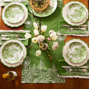 A table set for four features fern-patterned plates, green napkins, a green runner, white flowers in a vase, Caskata Library White Wine Glasses (set of 4), cutlery, and a bottle of wine.