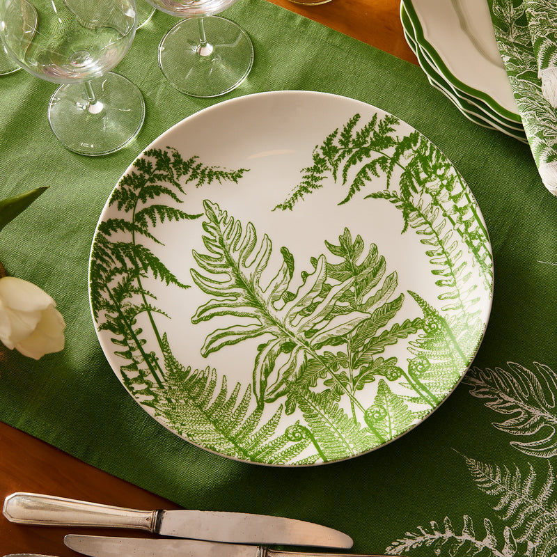 The Caskata Spring Verde Round Platter, featuring a green fern design, sits on a matching tablecloth, surrounded by glassware, cutlery, and stacked plates for an inviting tablescape.