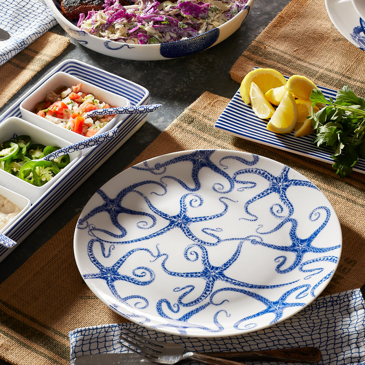 A table setting includes a Starfish Coupe Dinner Plate from Caskata Artisanal Home, a rectangular dish of lemon wedges and herbs, a bowl of salad, and a divided dish with various toppings, all on a burlap placemat. Adding to the charm are contemporary-shaped starfish accents subtly placed around the arrangement.