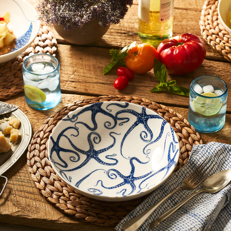 The Caskata Starfish Entrée Bowl, a ceramic dinnerware piece with blue octopus details, sits on a woven placemat amid drinks, food, and vegetables. Dishwasher safe, it adds ocean charm to any meal.