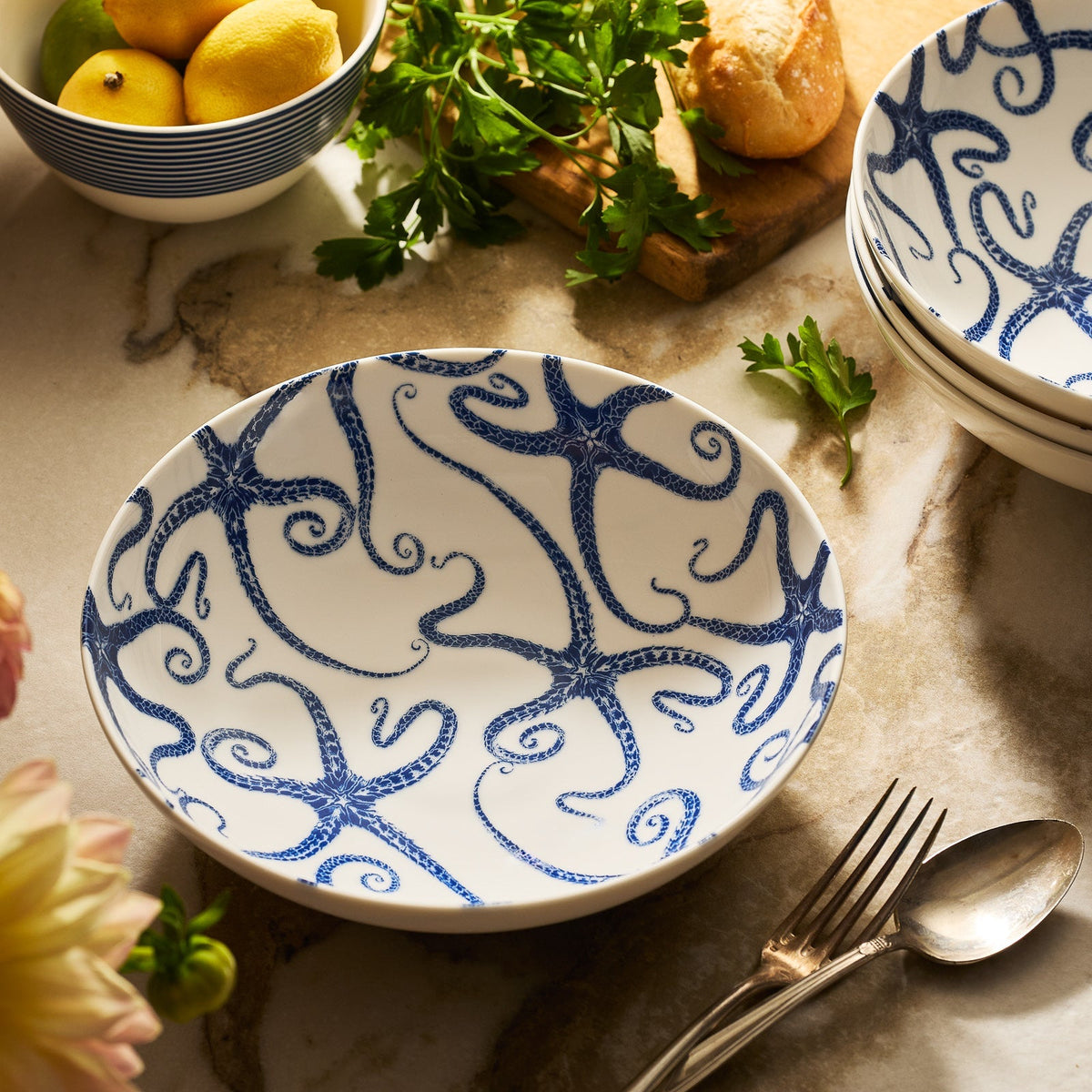 The Caskata Starfish Entrée Bowl, a dishwasher-safe ceramic with a blue starfish design, sits on a marble countertop beside a fork, spoon, parsley, lemons, and bread, bringing coastal charm to your kitchen.