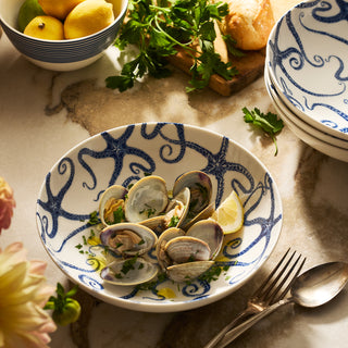 Cooked clams garnished with parsley and lemon, served in Caskata's Starfish Entrée Bowl on a marble table, accompanied by cutlery, bread, and fresh herbs.