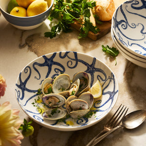 Cooked clams garnished with parsley and lemon, served in Caskata's Starfish Entrée Bowl on a marble table, accompanied by cutlery, bread, and fresh herbs.