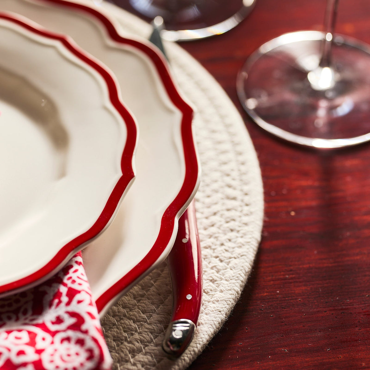 Two Caskata Stella Scalloped Crimson Dinner Plates are stacked on a woven placemat, paired with a red-handled knife, a patterned napkin, and a wine glass, all arranged on a wooden table.