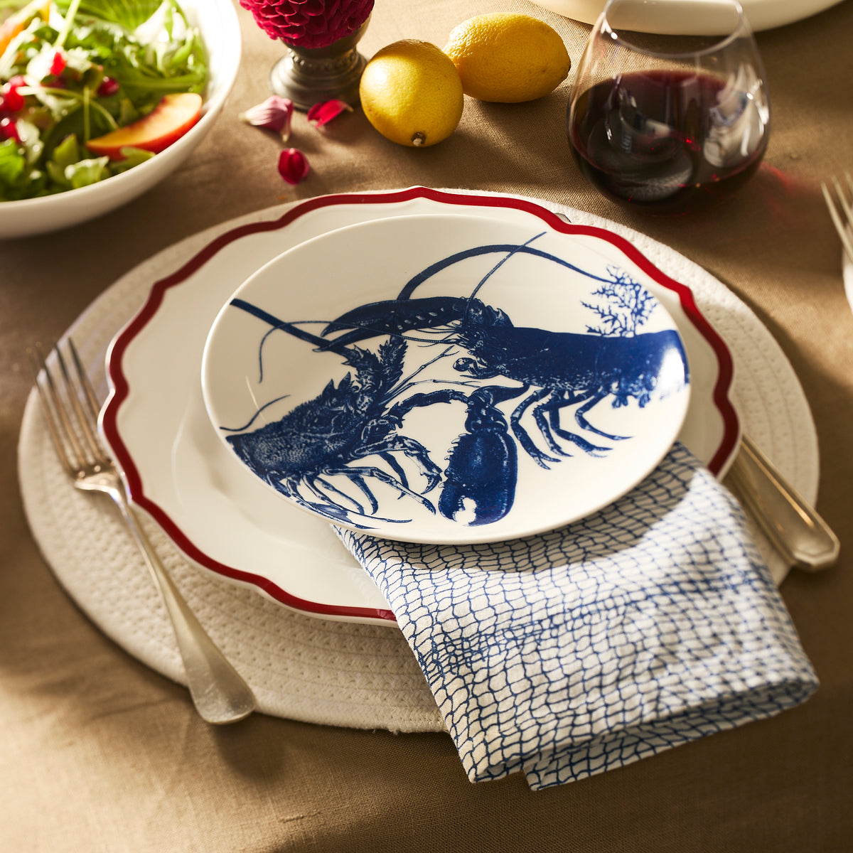 A dinner setting with a napkin, fork, knife, wine glass, salad, lemons, and a high-fired porcelain plate featuring a blue lobster illustration sits atop the Caskata Stella Scalloped Crimson Dinner Plate with a red rim.