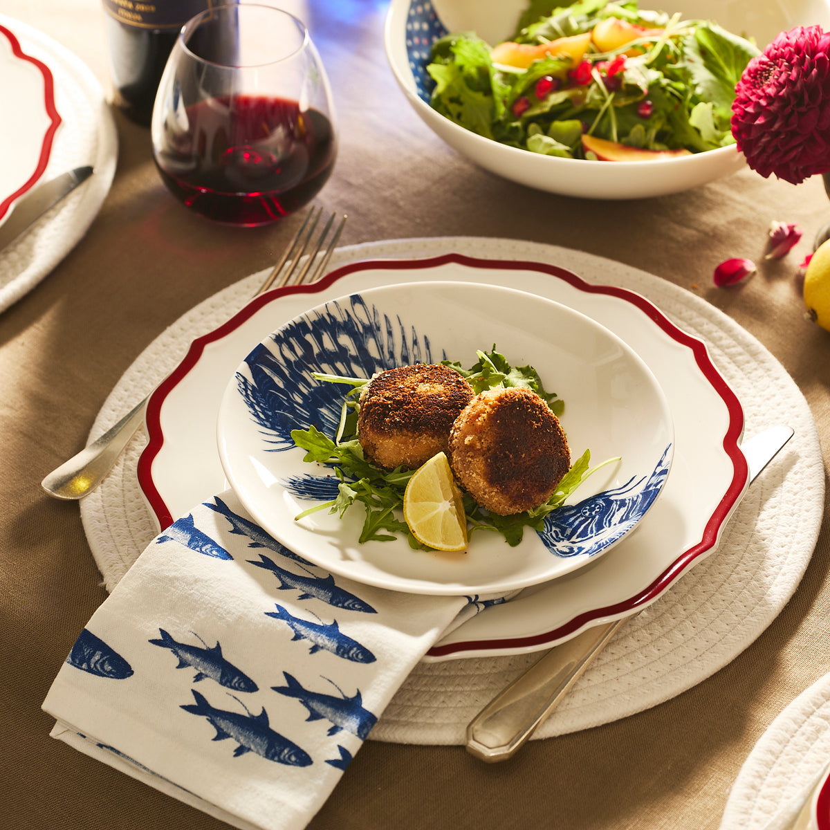 Two breaded crab cakes on greens with a lemon wedge, served on a Caskata Spiky Fish Coupe Salad Plate, beside a blue fish print napkin and a glass of red wine.