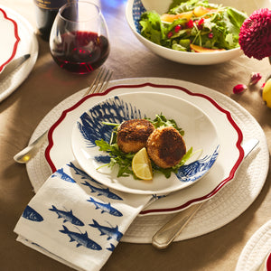 Two breaded crab cakes on greens with a lemon wedge, served on a Caskata Spiky Fish Coupe Salad Plate, beside a blue fish print napkin and a glass of red wine.
