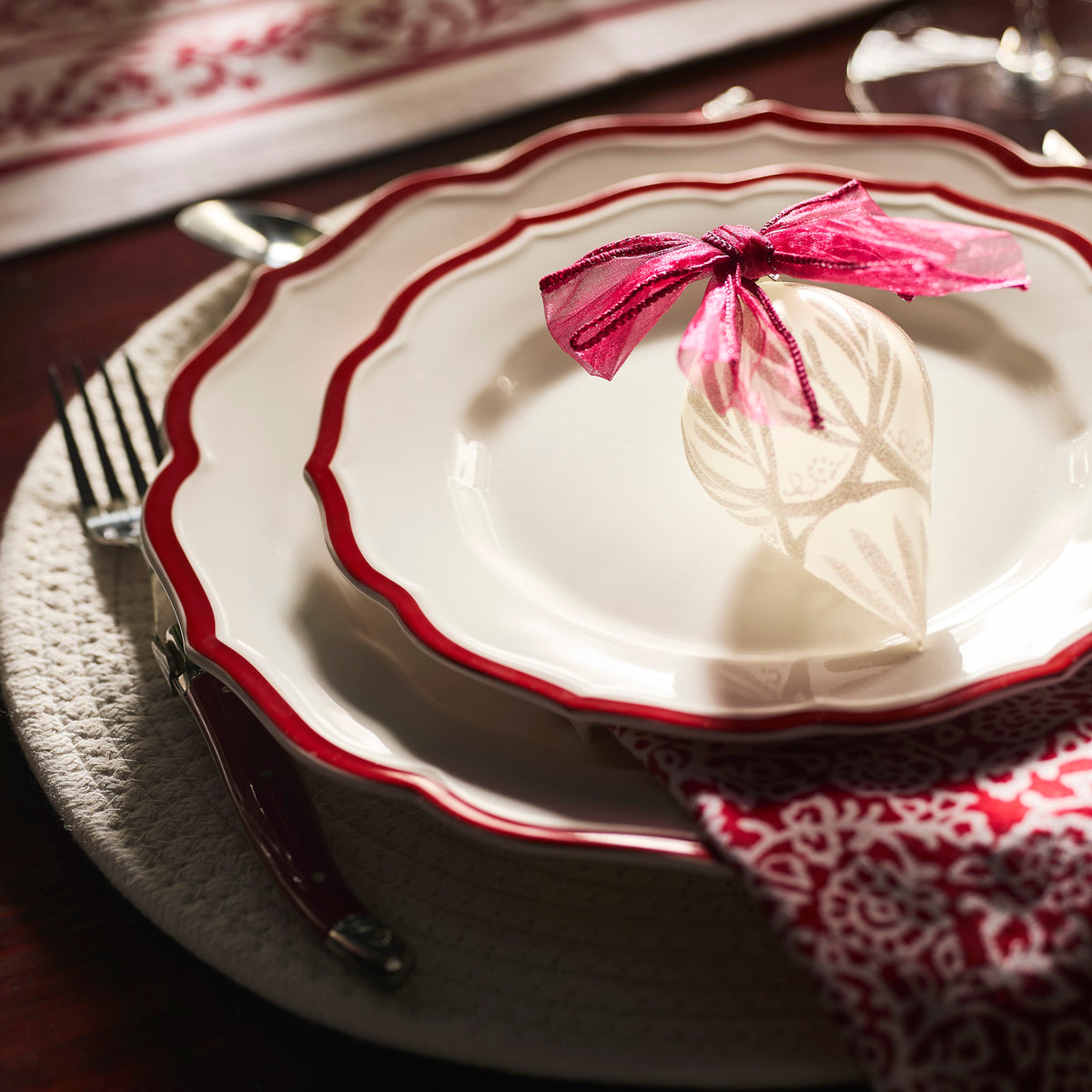 A close-up of a place setting featuring Caskata&#39;s Stella Scalloped Crimson Salad Plate, paired with a red-patterned napkin, silverware, and a decorative ornament tied with a pink ribbon on the plate.