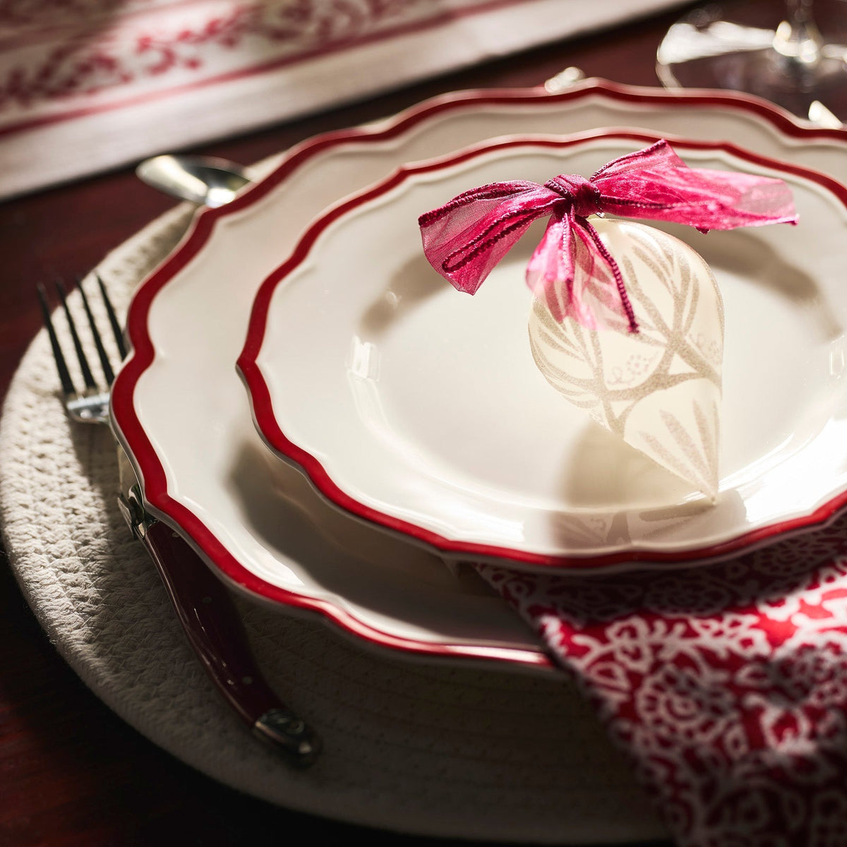 A close-up of a place setting featuring Caskata's Stella Scalloped Crimson Salad Plate, paired with a red-patterned napkin, silverware, and a decorative ornament tied with a pink ribbon on the plate.