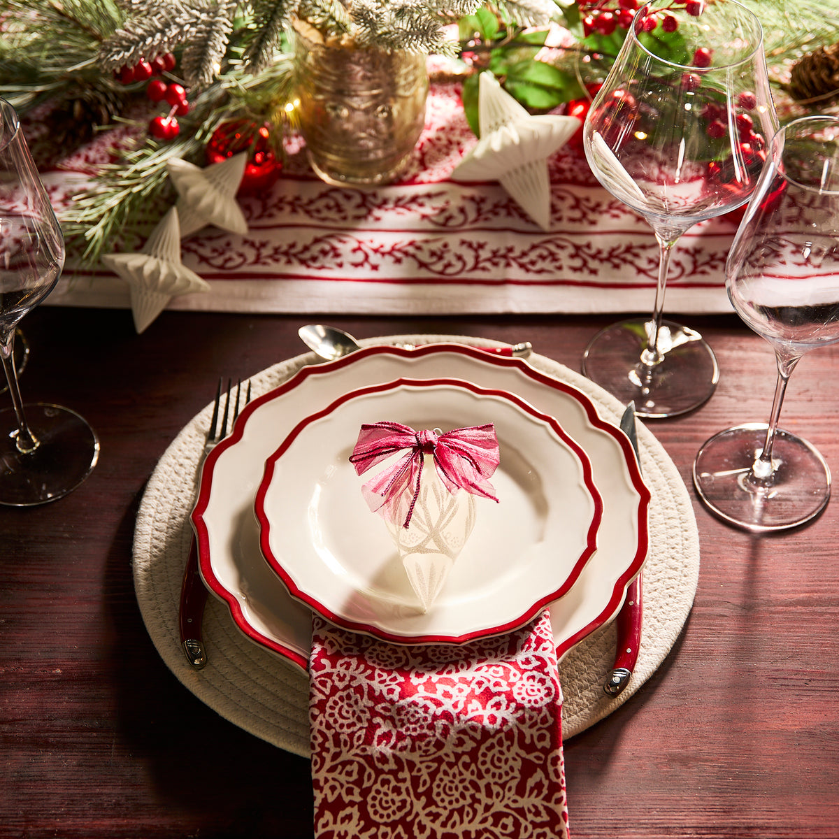 Create a festive holiday table with Caskata&#39;s Stella Scalloped Crimson Salad Plates, vintage-style dinnerware, patterned napkins, wine glasses, silverware, and decorative ornaments surrounded by Christmas greenery.