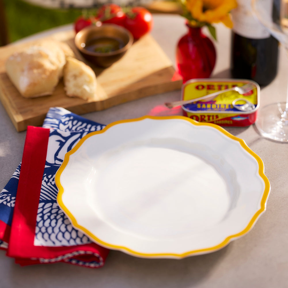 The Caskata Stella Scalloped Marigold Dinner Plate, featuring a yellow rim and white center, is set on the table beside a blue and red napkin, fresh bread, olive oil, tomatoes, canned sardines, and a wine glass for an inviting display.