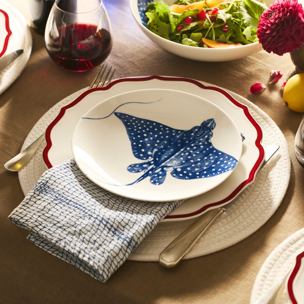 A dining table setting with a Caskata Stingray Coupe Salad Plate decorated with a blue illustration, accompanied by a napkin, cutlery, a glass of red wine, and a bowl of salad in the background.