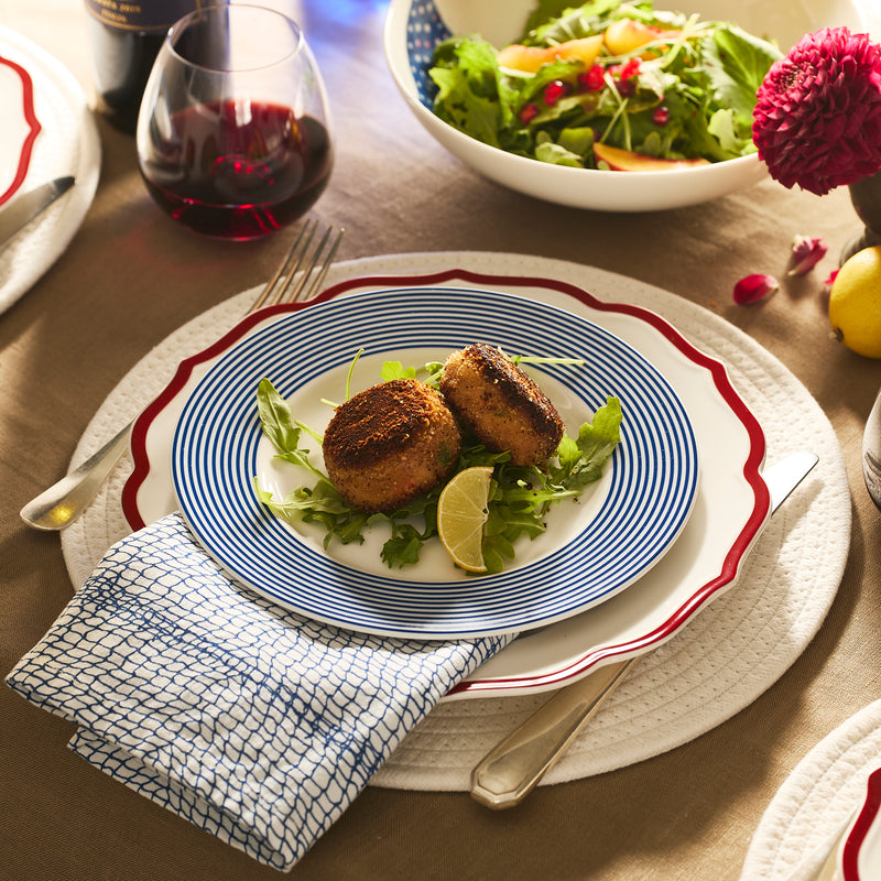 Two breaded patties on greens with lemon wedges, served on the Caskata Stella Scalloped Crimson Dinner Plate, accompanied by salad, red wine, and tableware on a neatly arranged dining table.