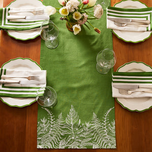 A wooden table set for four features green and white striped napkins, scalloped plates, clear glassware, and the Caskata Spring Verde Table Runner with a white fern motif. A flower vase in the center adds to the charming cottage-core style.