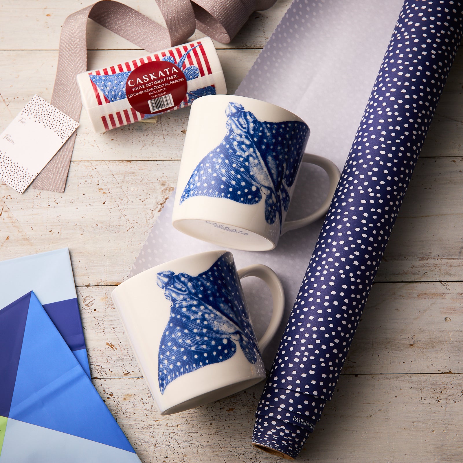 The "Stingray Coffee Talk" Bundle by Caskata features two porcelain mugs with blue manta ray designs, displayed on wood amid wrapping paper, ribbons, and colored sheets—perfect as a beach lover gift.