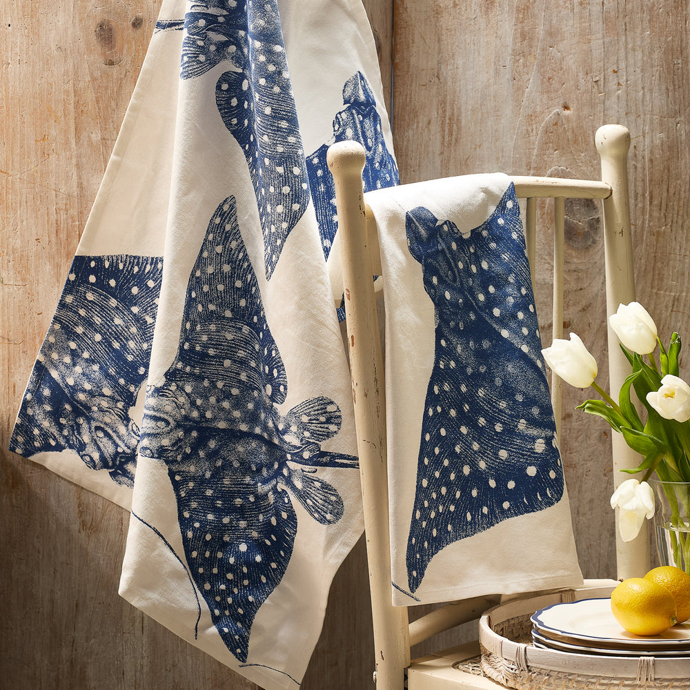 A cream chair displays Caskata's Stingray Kitchen Towels, Set of 2—cotton towels with stingray prints—adding a nautical accent beside a table with plates, a glass, lemons, and white tulips.