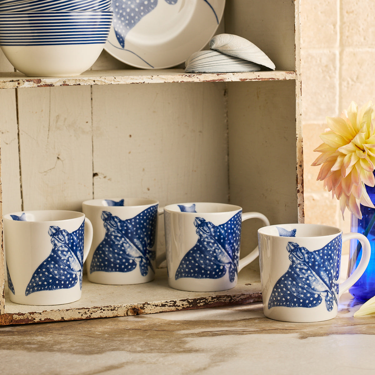 Four Caskata &quot;Ray&quot; the Stingray Mugs, featuring blue and white fish designs, are lined up on a counter by a rustic white shelf and a blue vase with a pink and yellow flower, creating a perfect coastal mug display.