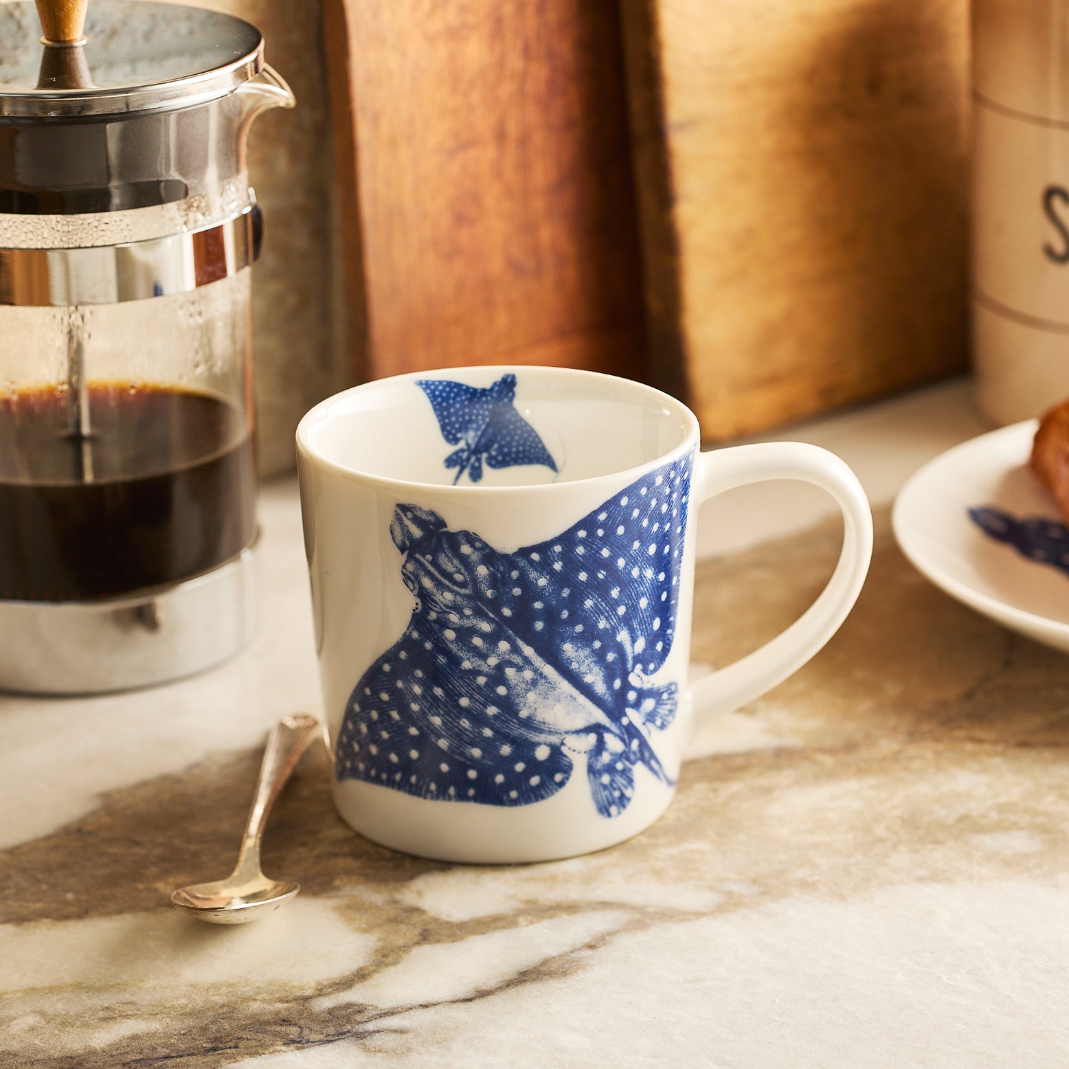 The "Stingray Coffee Talk" Bundle by Caskata features two porcelain mugs with blue manta ray designs, displayed on wood amid wrapping paper, ribbons, and colored sheets—perfect as a beach lover gift.