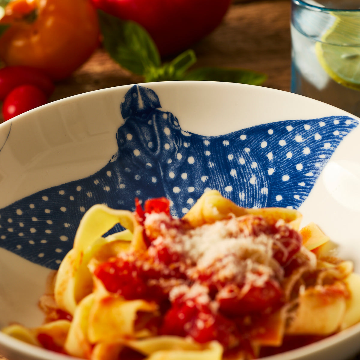 The &quot;Ray&quot; the Stingray Entrée Bowl by Caskata holds pasta with tomato sauce and cheese, while veggies and a lime water glass in the background enhance the coastal dinnerware charm.