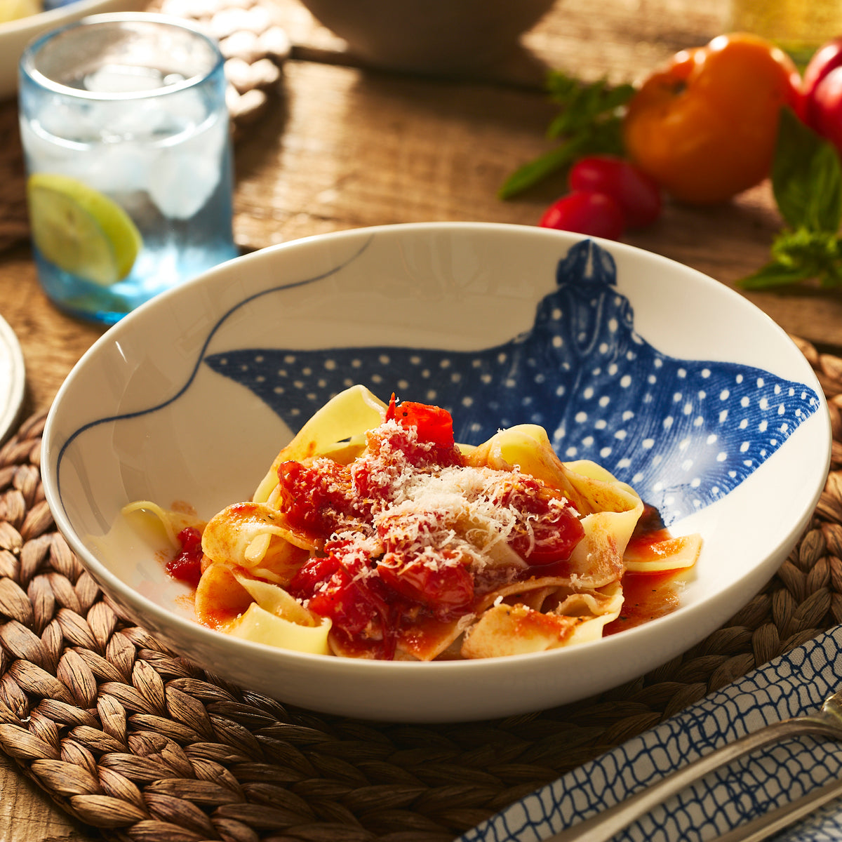 A bowl of pasta with tomato sauce and cheese is served in the Caskata &quot;Ray&quot; the Stingray Entrée Bowl, set on a woven placemat, with a glass of water and fresh vegetables in the background.