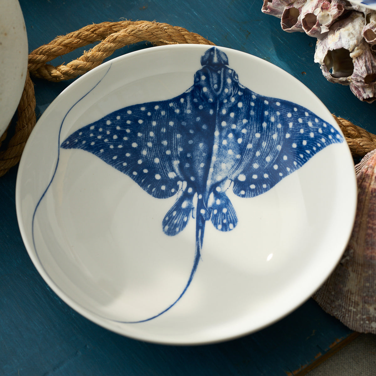 The "Ray" the Stingray Entrée Bowl by Caskata features a blue spotted stingray illustration at its center, shown on a blue surface with rope and seashells nearby.