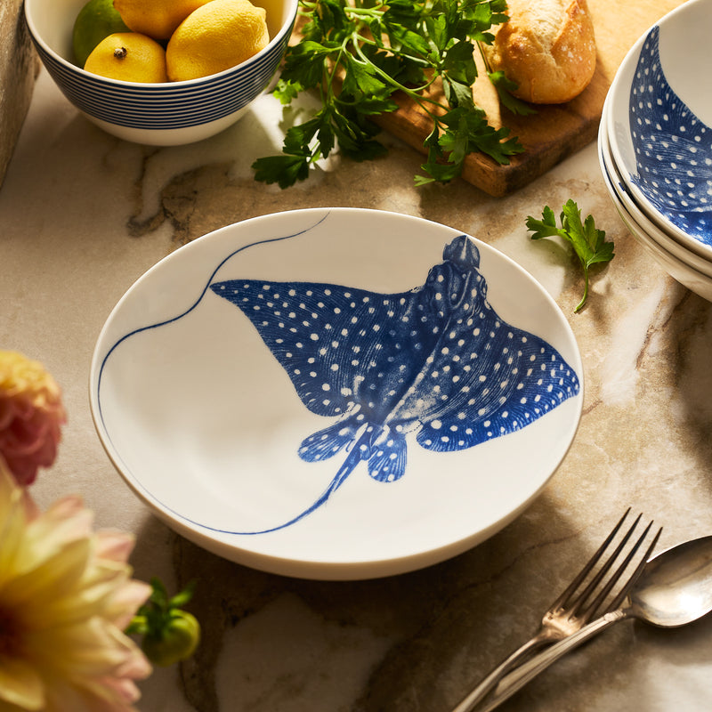 The "Ray" the Stingray Entrée Bowl by Caskata features a blue design and sits on marble near a fork, parsley, a bread roll, and lemons—perfect for adding coastal charm to your table setting.