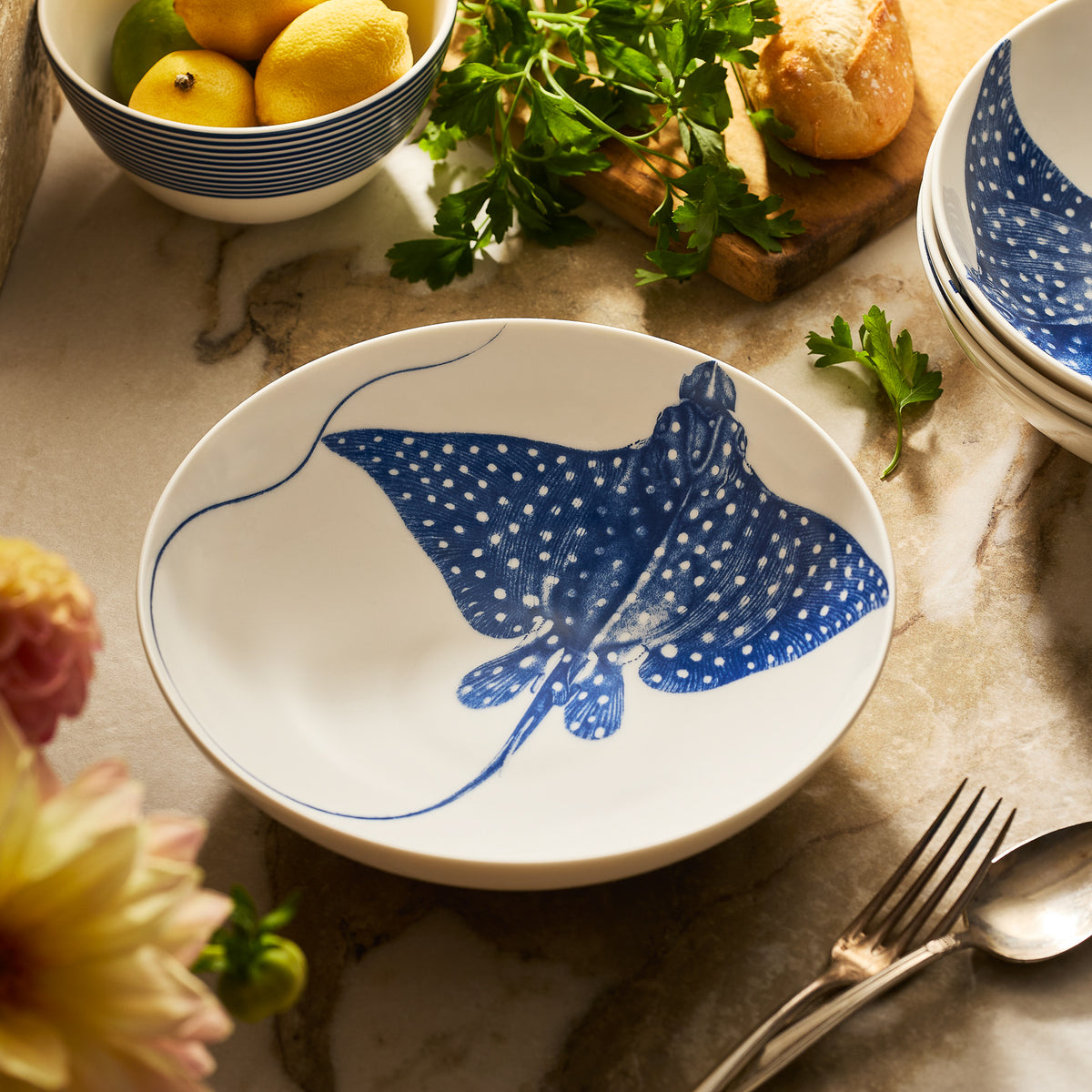 The &quot;Ray&quot; the Stingray Entrée Bowl by Caskata features a blue design and sits on marble near a fork, parsley, a bread roll, and lemons—perfect for adding coastal charm to your table setting.