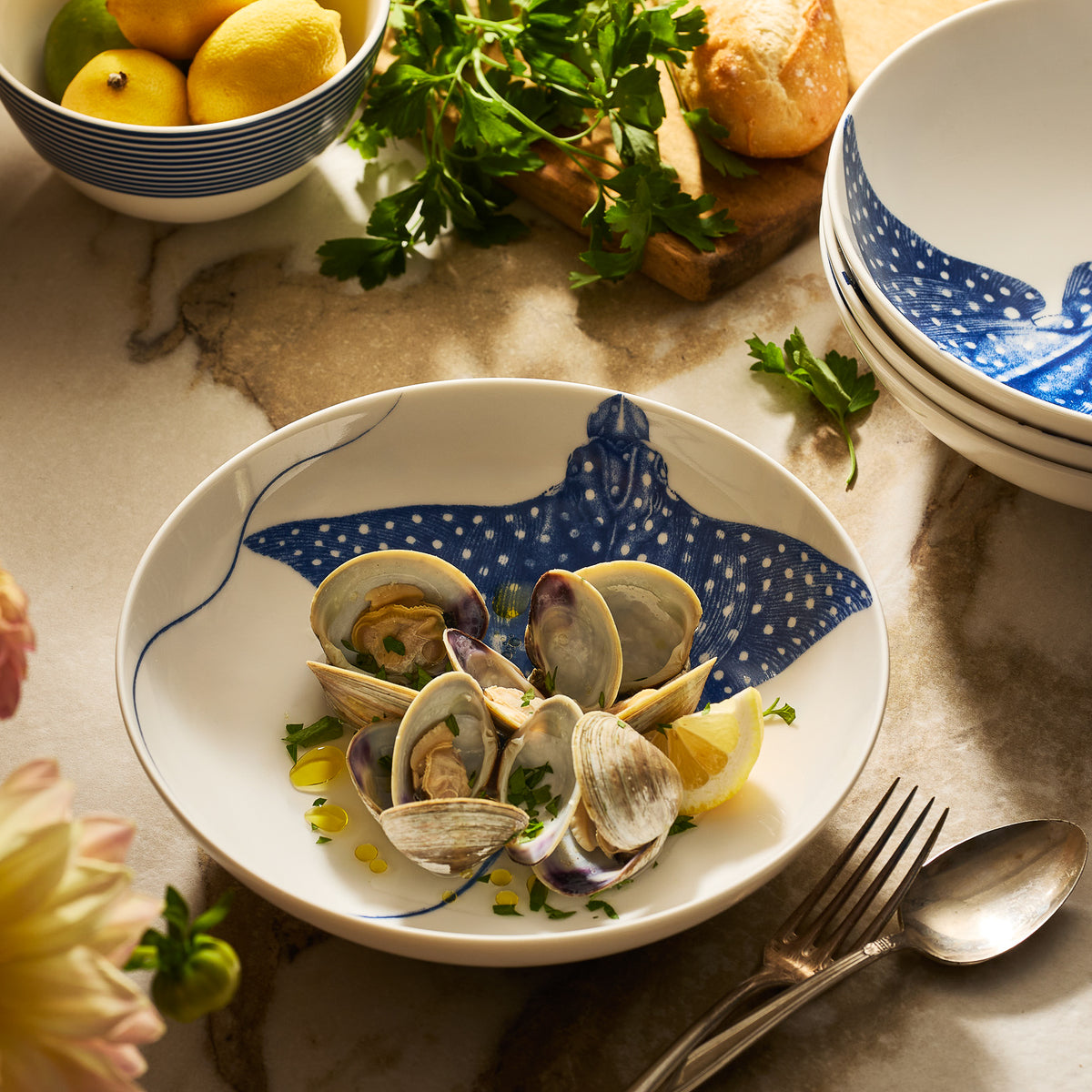 A plate of clams garnished with parsley and lemon rests on Caskata’s &quot;Ray&quot; the Stingray Entrée Bowl, surrounded by bread, utensils, and lemons—an elegant way to showcase coastal dinnerware.