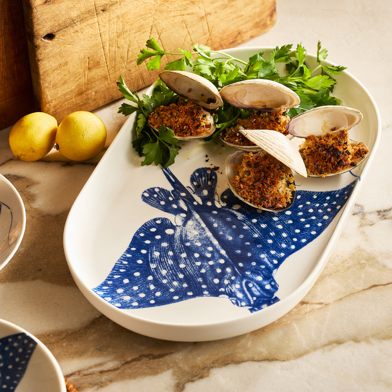 The Caskata "Ray" the Stingray Large Coupe Oval Platter from the Coastal Collection showcases baked stuffed clams on parsley, with two lemons and a wooden board in the background.