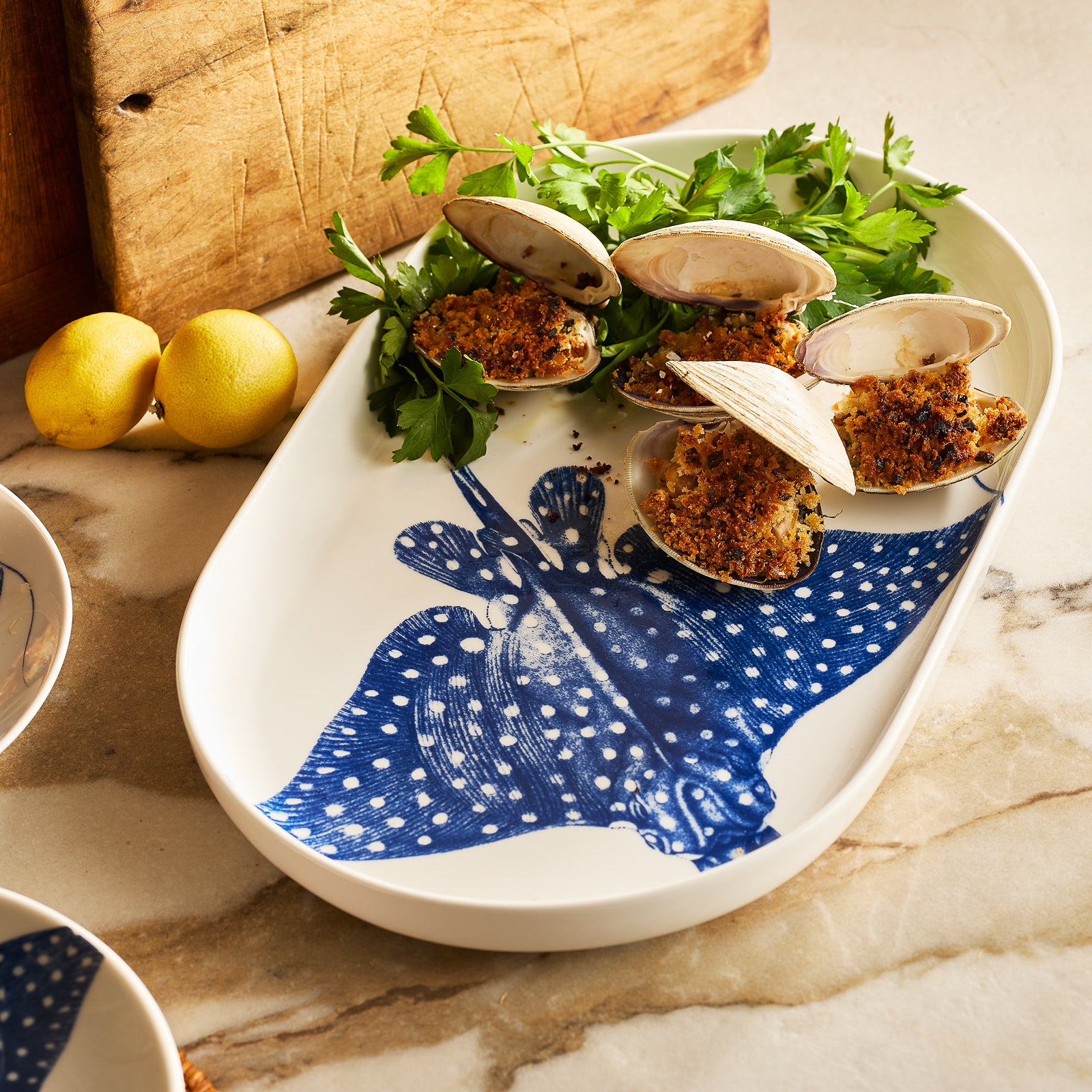 The "Ray" the Stingray Large Coupe Oval Platter by Caskata is a white porcelain piece from the coastal collection, featuring a blue illustration of a spotted eagle ray.