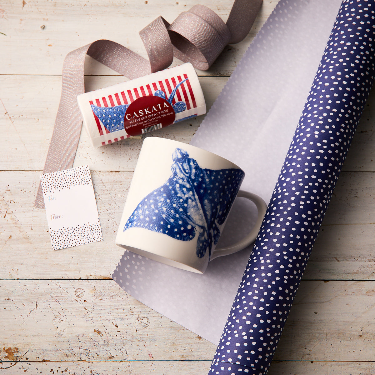 Caskata’s "Ray of Sunshine" Bundle, featuring a stingray mug with blue design, cocktail napkin roll, dotted wrapping paper, ribbon, gift tag, and red-and-white box, is displayed on light wood—perfect for an ideal gift set.