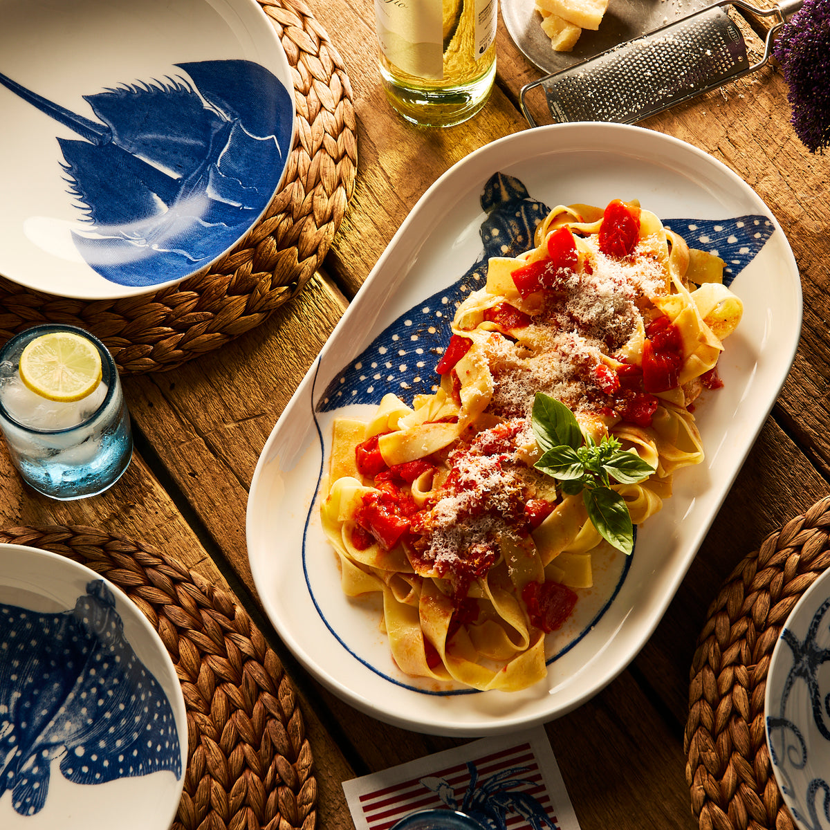 A platter of tagliatelle pasta with tomato sauce and grated cheese rests on a wooden table, surrounded by blue and white fish-patterned plates, woven placemats, and a Caskata Horseshoe Crab Entrée Bowl.