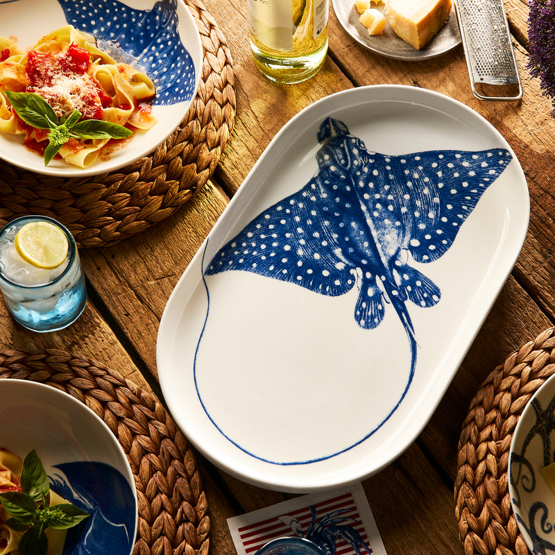 The "Ray" the Stingray Large Coupe Oval Platter by Caskata from the Coastal Collection rests on a wooden table set for dining, surrounded by plates of pasta, drinks, and woven placemats.