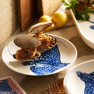 Two baked stuffed clams sit on a Caskata Stingray Coupe Salad Plate, featuring a blue pattern, with lemons and greens in the background on a marble countertop.