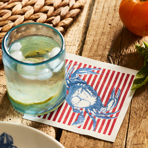 A glass of iced water rests on the MY DRAP Seaside Stripes Cocktail Napkin Roll atop a wooden table, beside a woven placemat, fresh basil leaves, and an orange tomato.