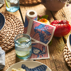 The MY DRAP Seaside Stripes Cocktail Napkin Roll, with blue crab prints and red stripes, brightens your table with coastal charm beside a glass of water, a tomato, and woven placemats.