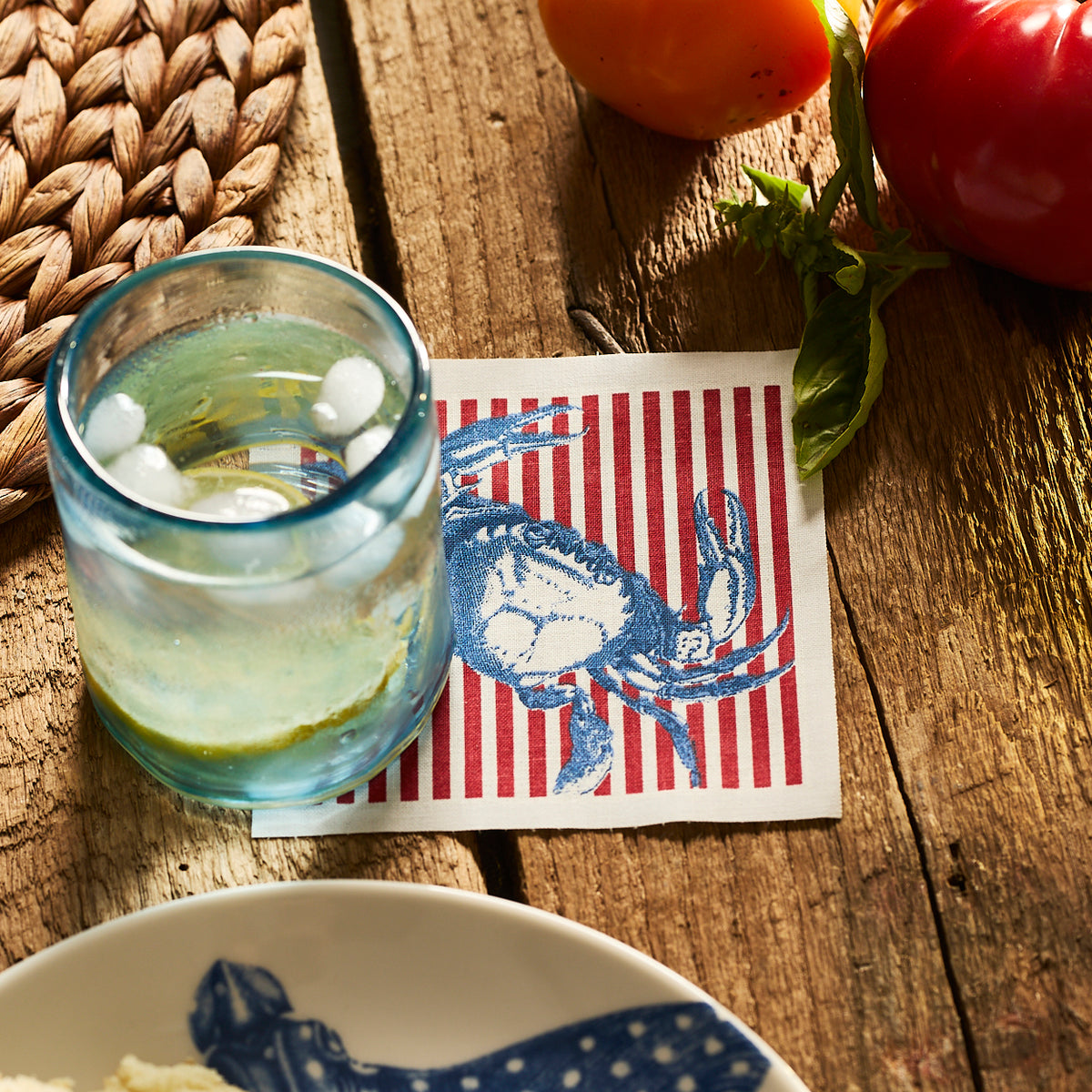 A blue glass with ice rests on the MY DRAP Seaside Stripes Cocktail Napkin Roll, featuring a blue crab, next to tomatoes and a woven placemat on a wooden table.