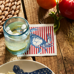 A blue glass with ice rests on the MY DRAP Seaside Stripes Cocktail Napkin Roll, featuring a blue crab, next to tomatoes and a woven placemat on a wooden table.