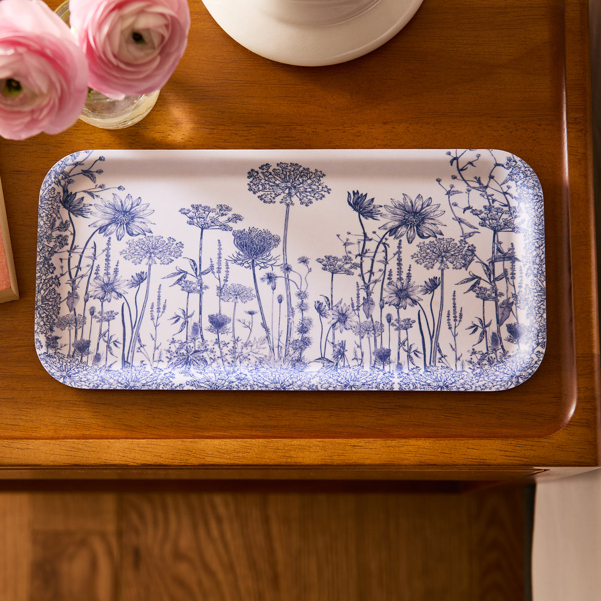 A Caskata Summer Blues Small Rectangular Birchwood Tray, featuring blue floral patterns on white, sits on wood with pink flowers nearby, highlighting its eco-friendly design.