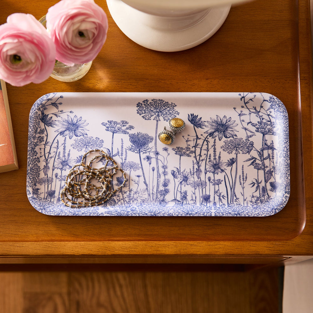 The Caskata Summer Blues Small Rectangular Birchwood Tray, made from sustainable birchwood, elegantly displays a chain necklace and gold earrings on a wooden table adorned with vibrant pink ranunculus flowers.