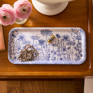 The Caskata Summer Blues Small Rectangular Birchwood Tray, made from sustainable birchwood, elegantly displays a chain necklace and gold earrings on a wooden table adorned with vibrant pink ranunculus flowers.