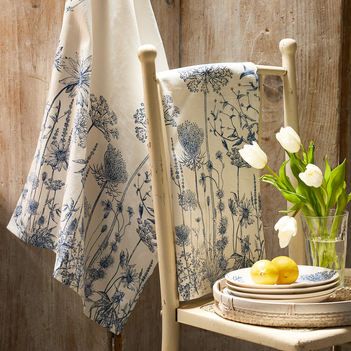 A chair with a white cloth featuring blue botanicals sits beside a table of tulips and lemons, perfectly complemented by Caskata's Summer Blues Kitchen Towels (Set of 2) against a rustic wooden background.