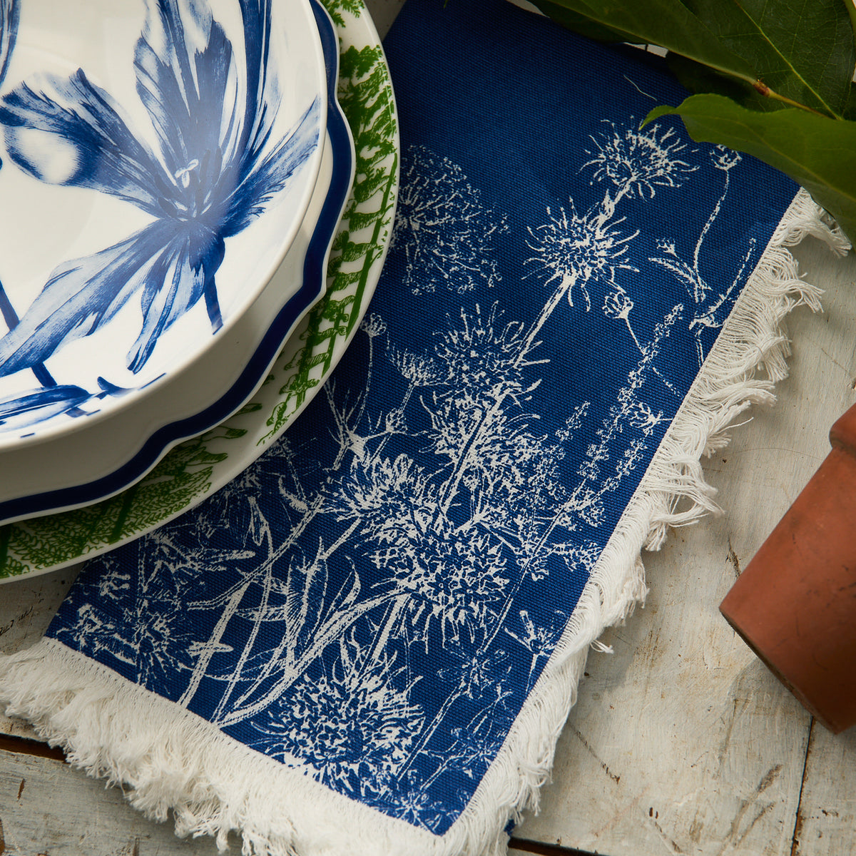 The Caskata Summer Blues Dinner Napkin, featuring white botanical prints on blue cotton, is displayed on wood beside patterned plates and a terracotta pot—an elegant cottage-core touch for your table linens.