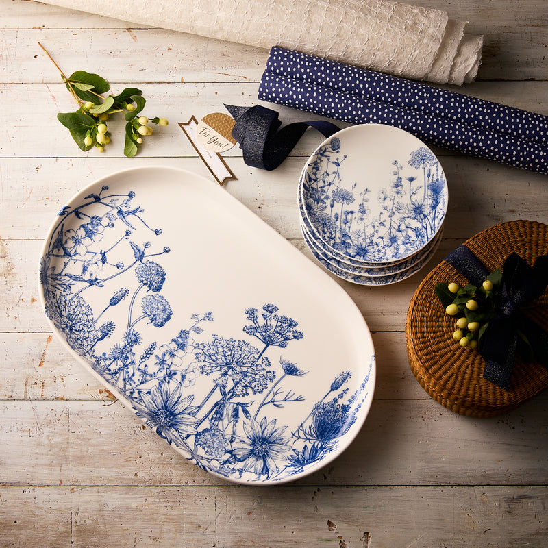 The "Wildflower Wonder" Bundle by Caskata features white oval and round appetizer plates with blue botanical designs, inspired by New England wildflowers, beautifully arranged on rustic wood alongside fabric rolls and greenery.