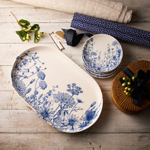 The Caskata Wildflower Wonder Bundle, a set of white porcelain serveware plates with blue floral designs inspired by New England wildflowers, is arranged on a rustic wooden surface with fabric, greenery, and a wicker basket.